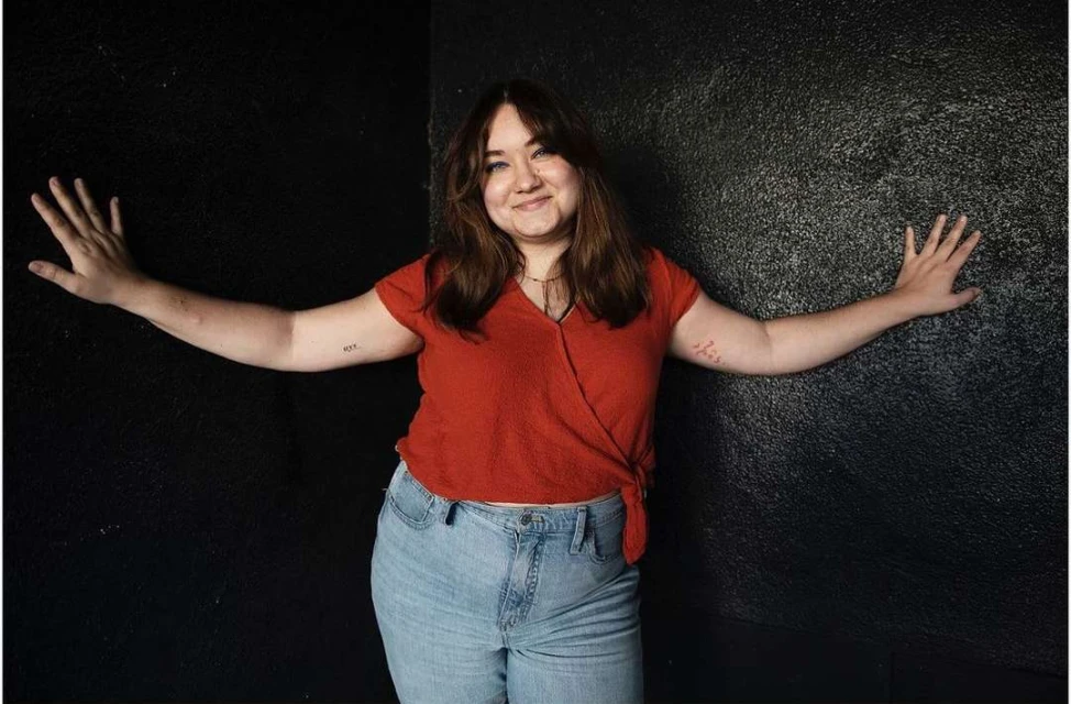 Portrait of a smiling young woman with her arms outstretched against a black wall, wearing a red shirt and light blue jeans.