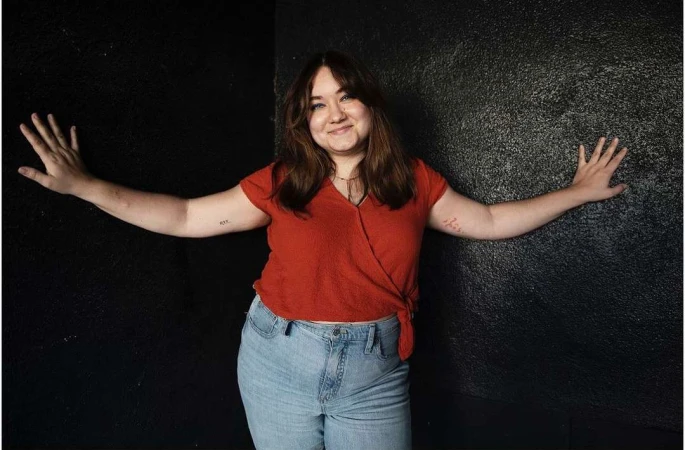 Portrait of a smiling young woman with her arms outstretched against a black wall, wearing a red shirt and light blue jeans.
