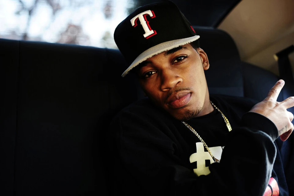 Young adult male in black top and baseball cap poses in a car, peace sign, gold chain, window background.
