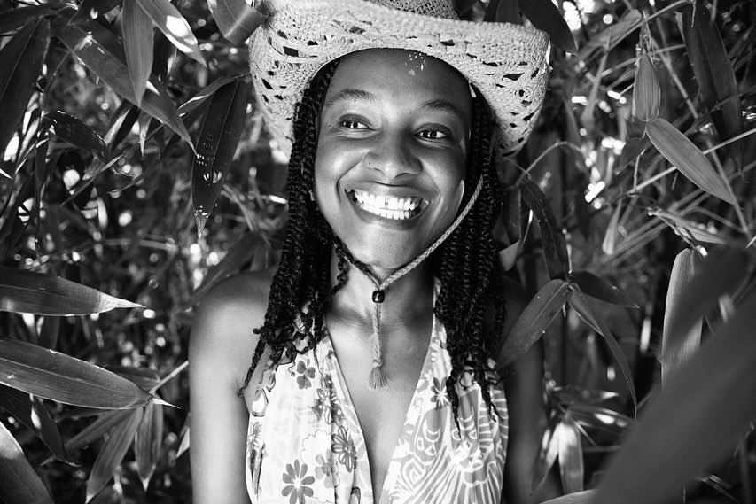 Black woman wearing a straw hat and patterned top smiling candidly amidst dense bamboo leaves in a monochrome portrait.