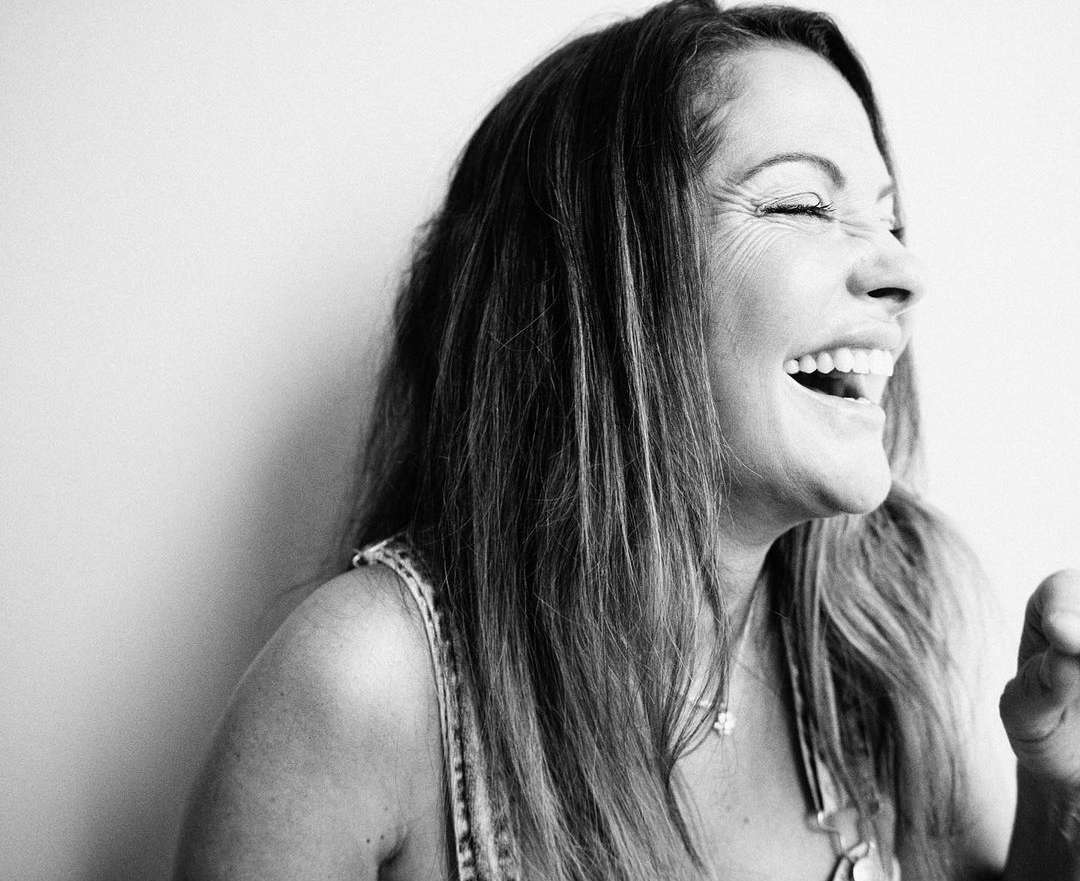 Close-up in black and white of an adult female smiling widely, with long hair and a sleeveless top against a plain background.