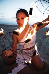 Woman in white one-shoulder outfit kneeling on sand holding a flaming fire prop with trails of fire against an ocean backdrop