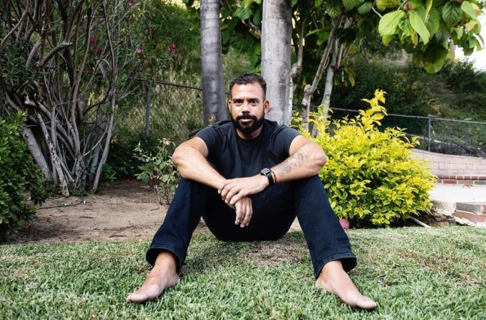 Man with beard sitting on green grass outdoors wearing a black t-shirt and dark trousers, photographed by Jeff Forney.