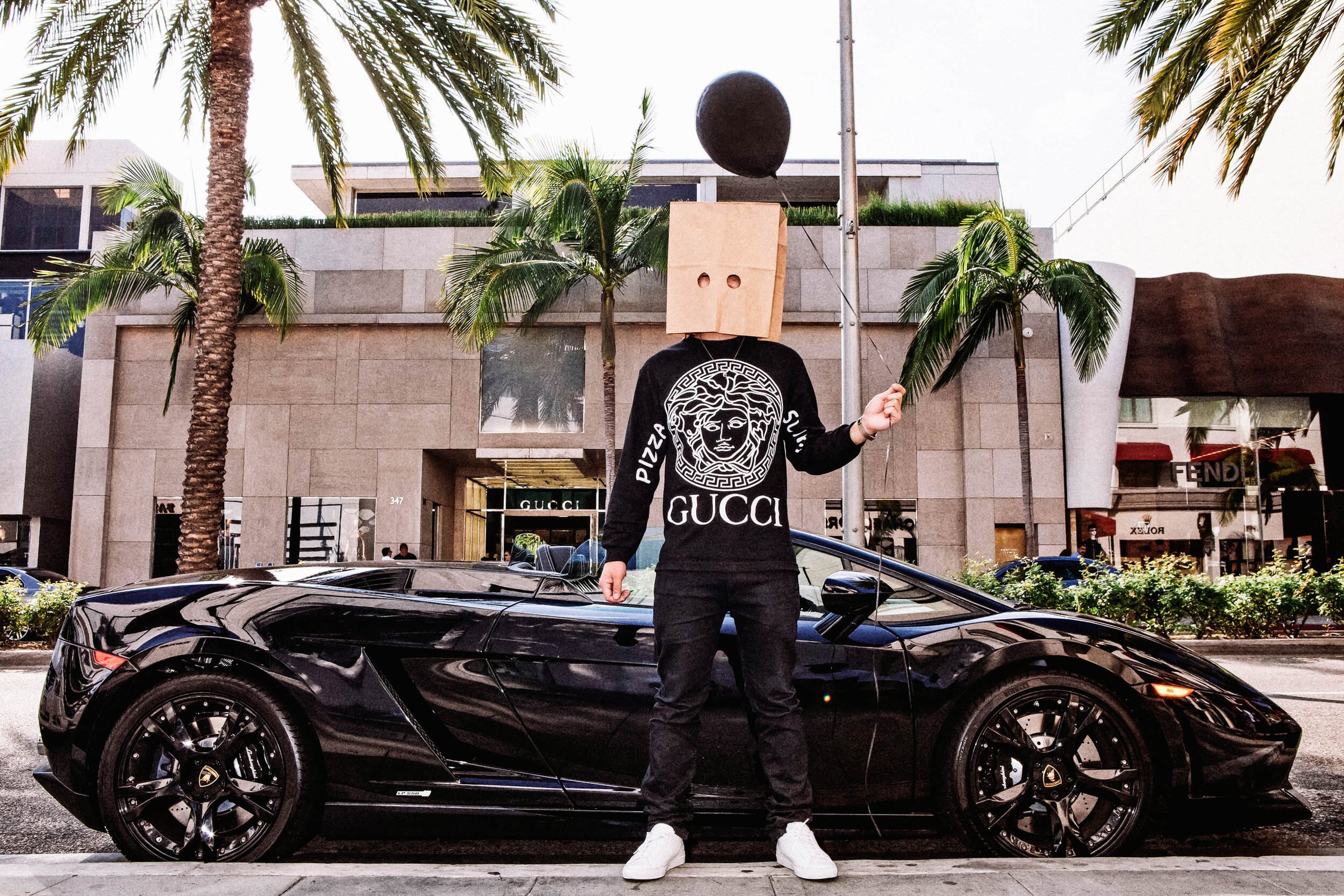Person in black Versace sweatshirt, black pants, white shoes, paper bag over head, holding balloon, next to black Lamborghini on street.