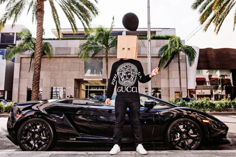 Person in black Versace sweatshirt, black pants, white shoes, paper bag over head, holding balloon, next to black Lamborghini on street.