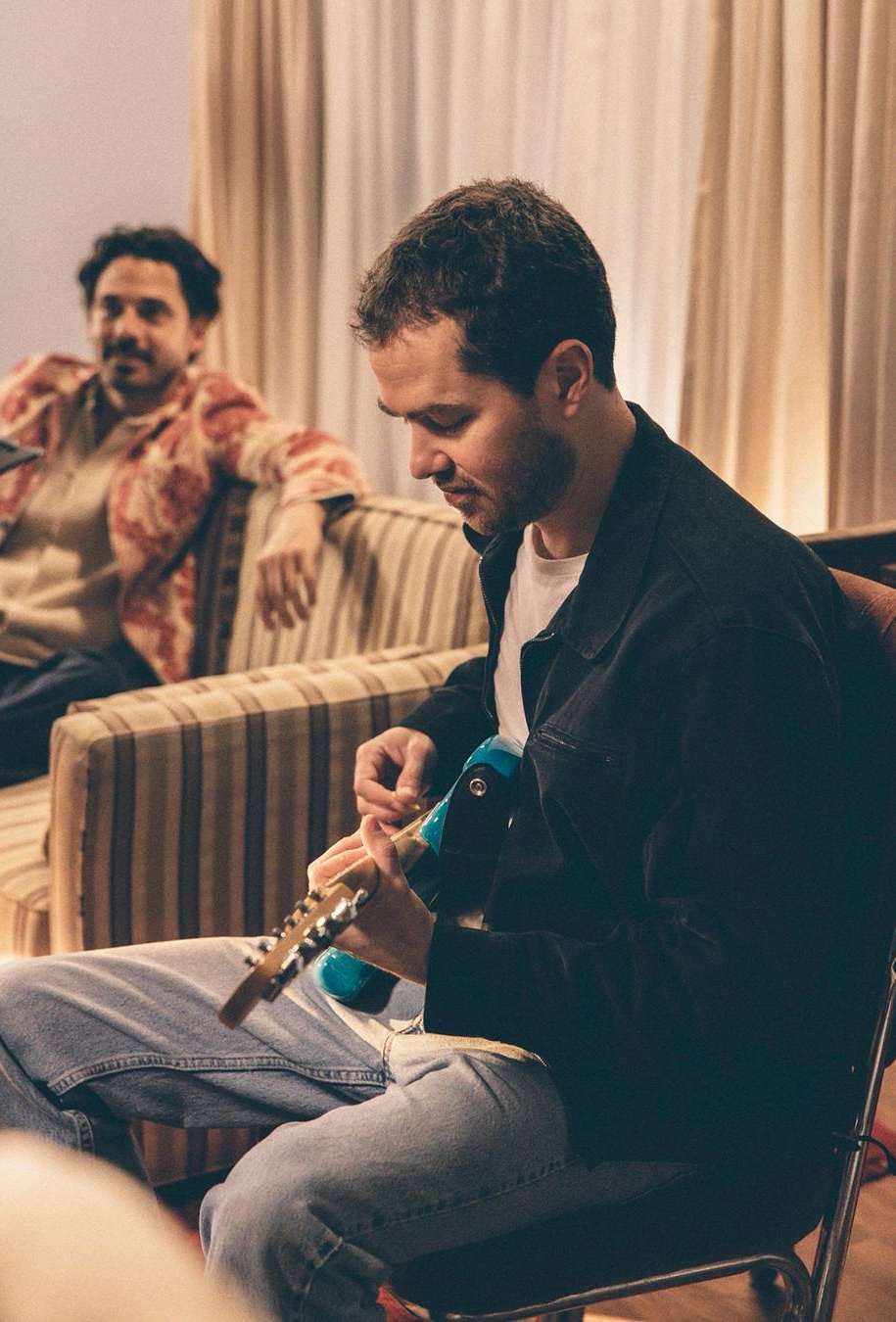 Two men in an indoor setting. One man plays a guitar while the other relaxes on a striped couch, both under soft, diffused light.