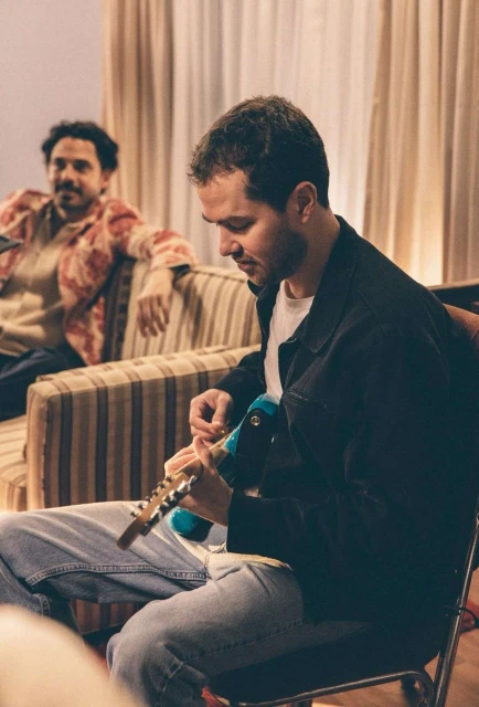 Two men in an indoor setting. One man plays a guitar while the other relaxes on a striped couch, both under soft, diffused light.