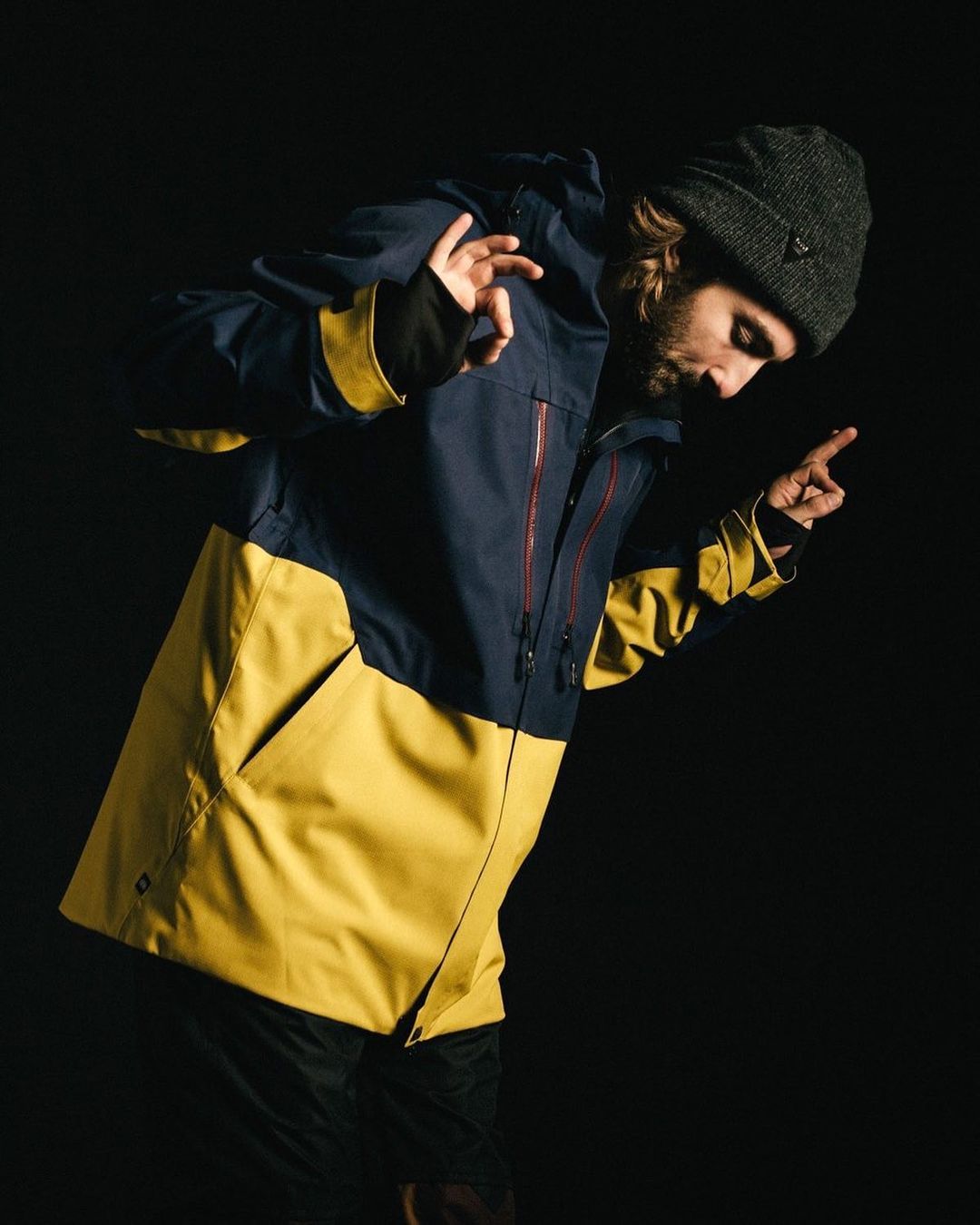 Man in a navy and yellow jacket and beanie poses indoors with a dark background, showcasing a casual and stylish outdoor look.