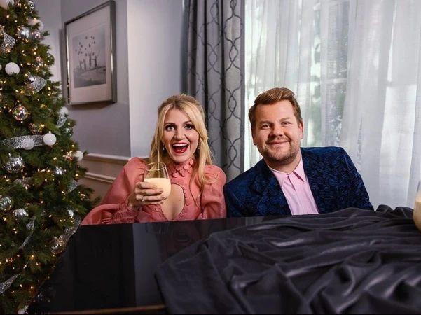 Festive indoor scene featuring a woman holding a glass of eggnog and a man in a blue jacket near a decorated Christmas tree.