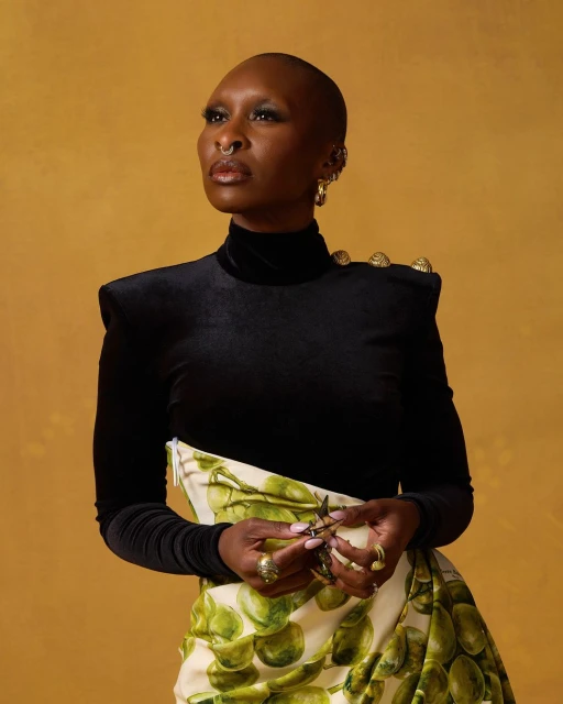 Woman with shaved head in black turtleneck and patterned skirt poses in a studio setting with a yellow background and gold jewelry.