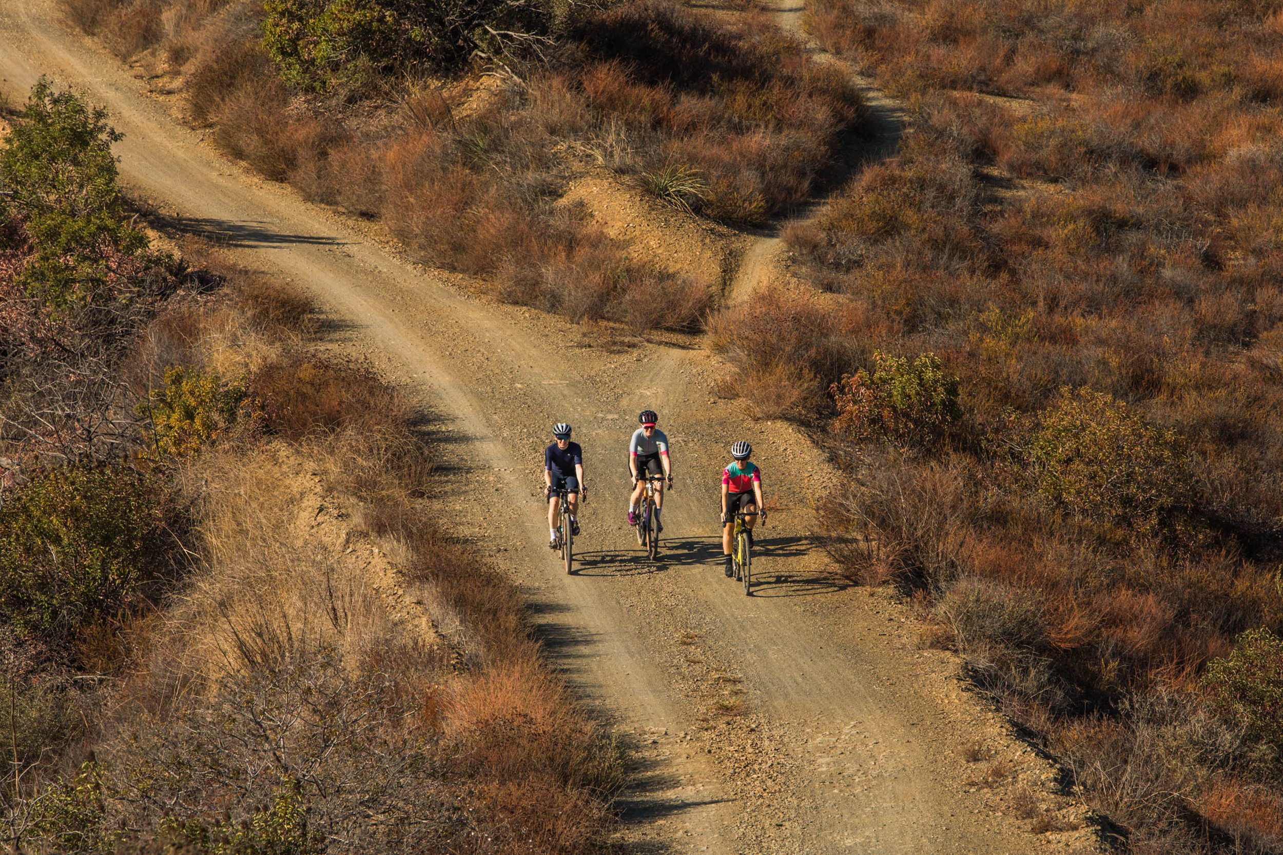 Three cyclists ride on a dirt road through a dry, shrub-filled landscape, captured from an elevated perspective under bright daylight.