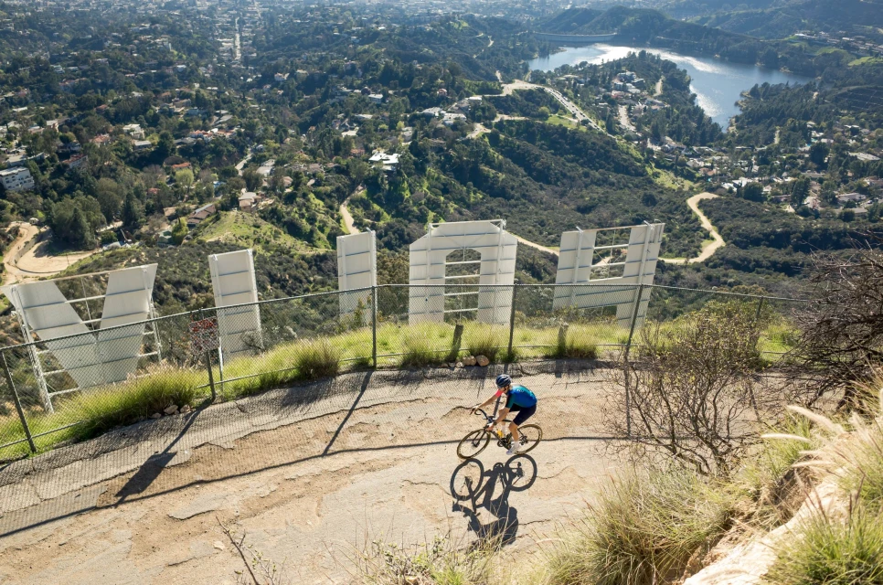 A cyclist rides along a dirt path near the Hollywood sign in Los Angeles with the city and a lake visible in the background.