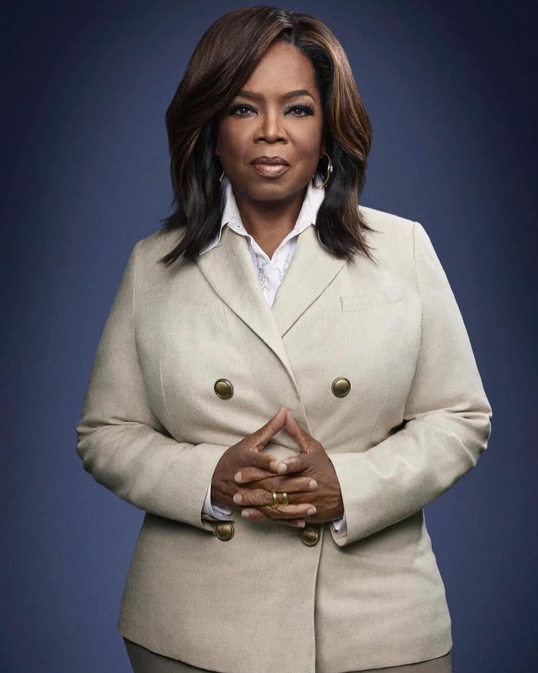 Headshot of Oprah Winfrey wearing a beige double-breasted blazer, facing forward with hands clasped, set against a dark blue background.