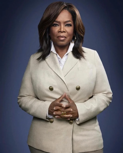 Headshot of Oprah Winfrey wearing a beige double-breasted blazer, facing forward with hands clasped, set against a dark blue background.