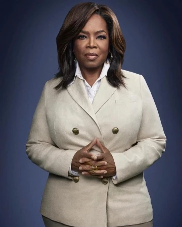 Headshot of Oprah Winfrey wearing a beige double-breasted blazer, facing forward with hands clasped, set against a dark blue background.