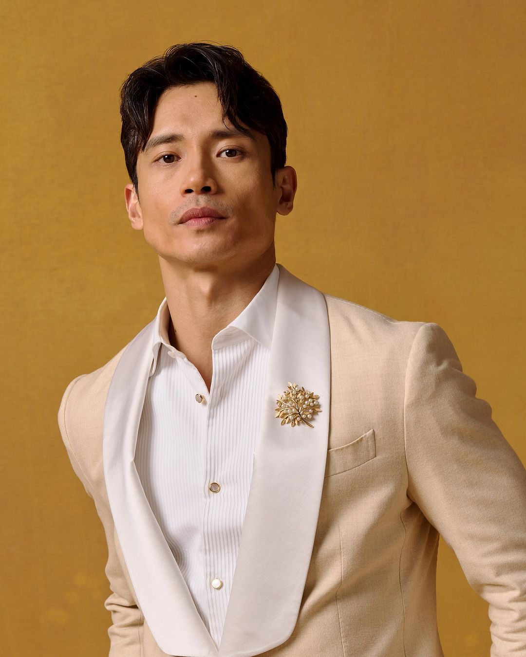 Headshot of a handsome adult male wearing a cream-colored suit with a white dress shirt against a yellow backdrop in a studio setting.