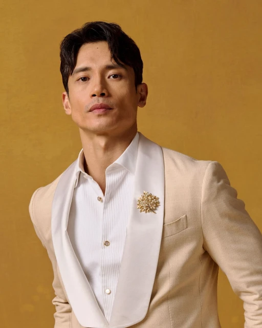 Headshot of a handsome adult male wearing a cream-colored suit with a white dress shirt against a yellow backdrop in a studio setting.