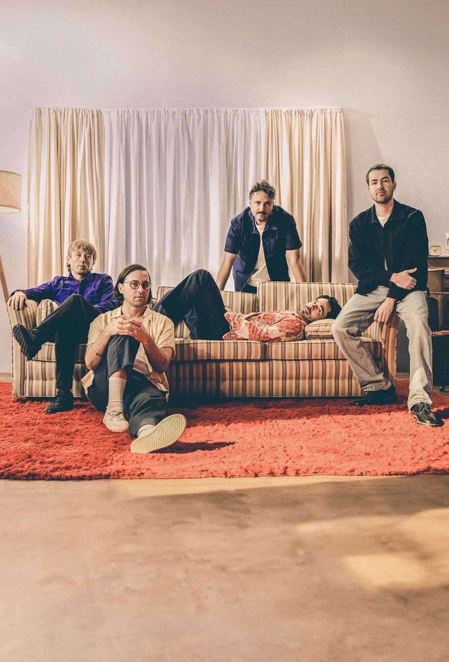 Group of five adults indoors: three seated, one lying on a striped couch, one leaning over the couch. All are casually dressed in a room.