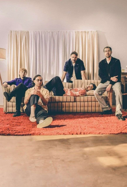 Group of five adults indoors: three seated, one lying on a striped couch, one leaning over the couch. All are casually dressed in a room.