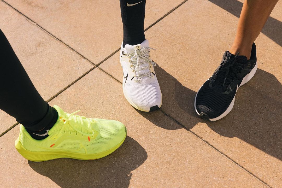 Three people wear various Nike running shoes standing on a concrete surface, promoting sports, fitness, and athletic wear in outdoor setting.