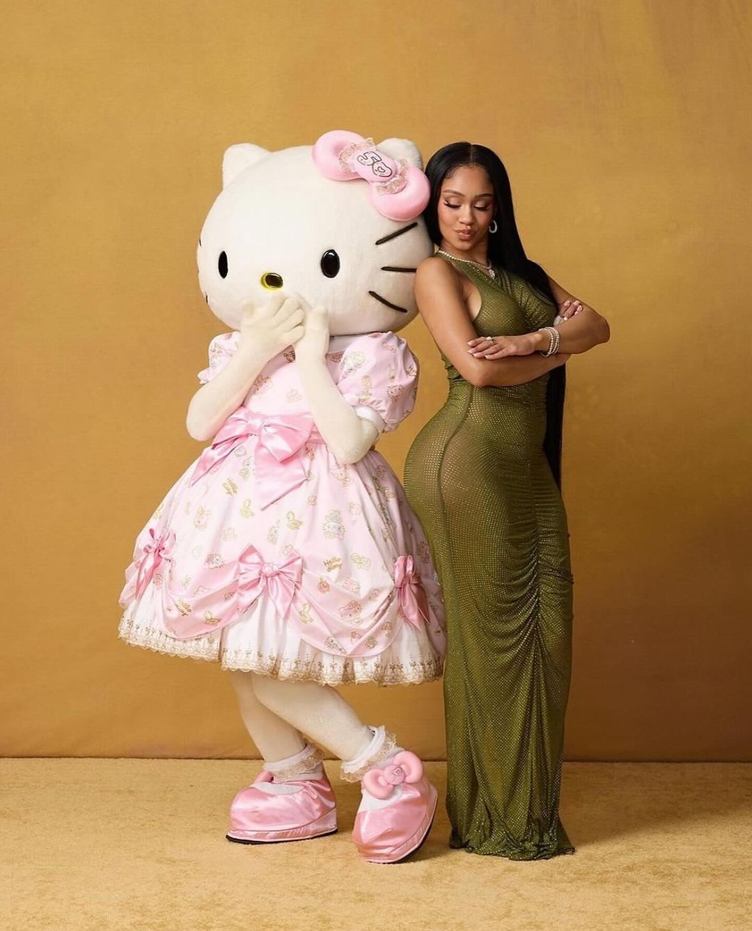 Adult woman in long green dress poses next to Hello Kitty mascot in pink dress against neutral backdrop, showcasing fashion and character collaboration.