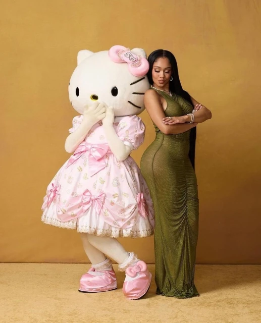 Adult woman in long green dress poses next to Hello Kitty mascot in pink dress against neutral backdrop, showcasing fashion and character collaboration.