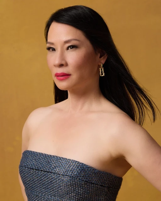 Close-up portrait of Lucy Liu with dark hair and an off-shoulder blue dress against a gold background, showcasing her elegant style.