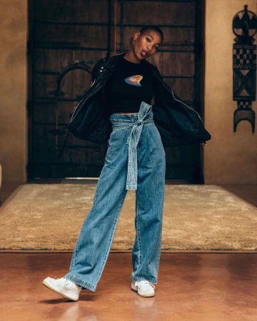 Young Black woman with a shaved head wearing high-waisted belted denim trousers, a black t-shirt, and a black jacket, posing in a room.