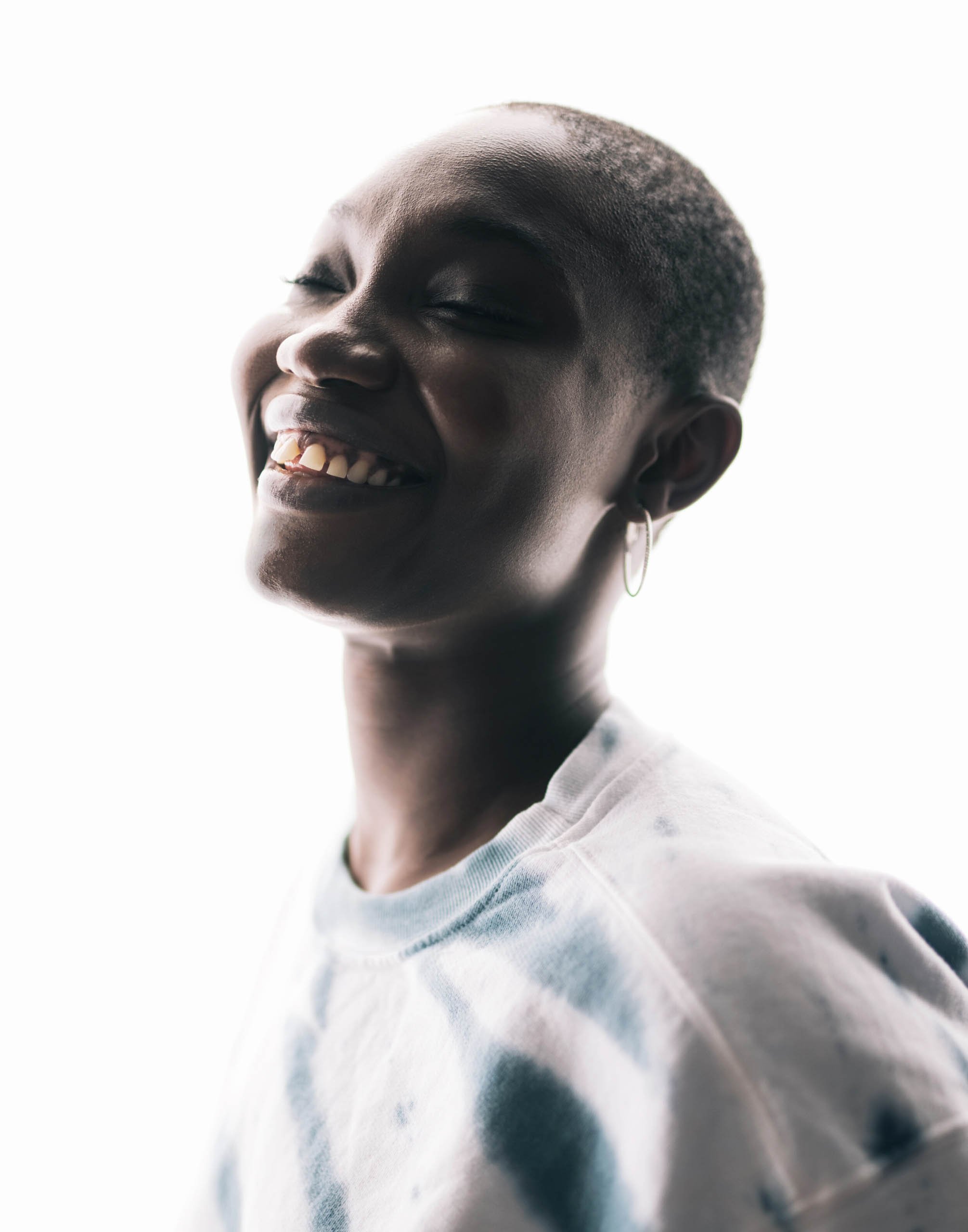 Portrait of a smiling dark-skinned woman with short hair, wearing a tie-dye top, against a bright white background. Eye-level angle, indoor lighting.