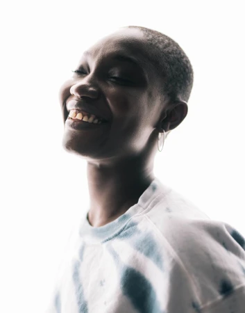 Portrait of a smiling dark-skinned woman with short hair, wearing a tie-dye top, against a bright white background. Eye-level angle, indoor lighting.