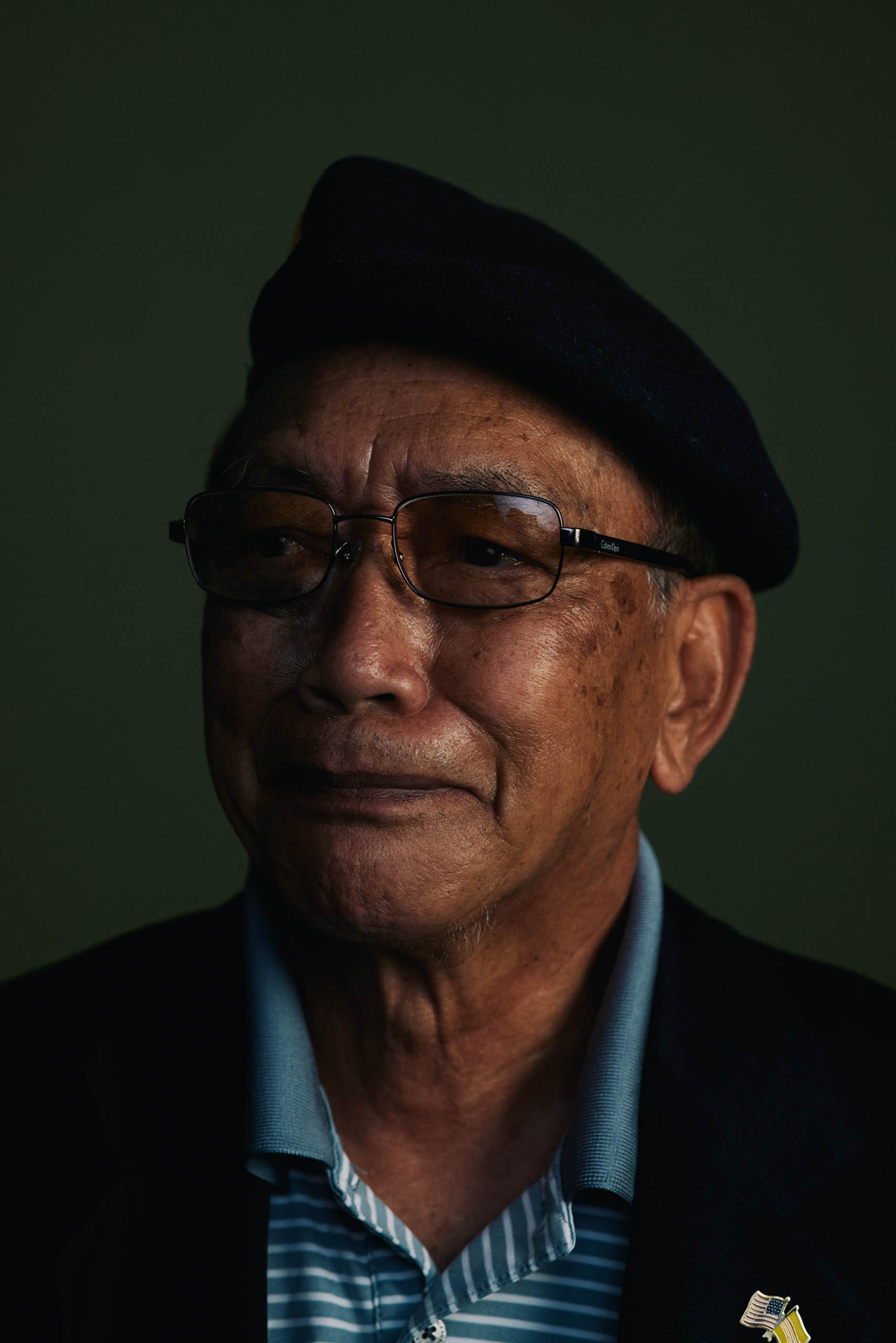 Elderly man wearing glasses, a dark beret, striped polo shirt, and dark jacket against a dark green studio backdrop.