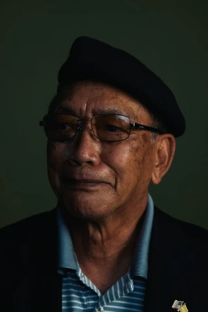Elderly man wearing glasses, a dark beret, striped polo shirt, and dark jacket against a dark green studio backdrop.