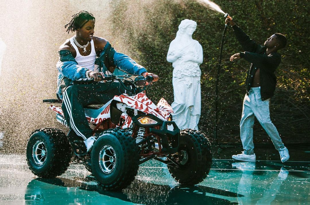 Young adult rides ATV as another uses hose to spray water, with statue and trees in background under natural lighting outdoors.