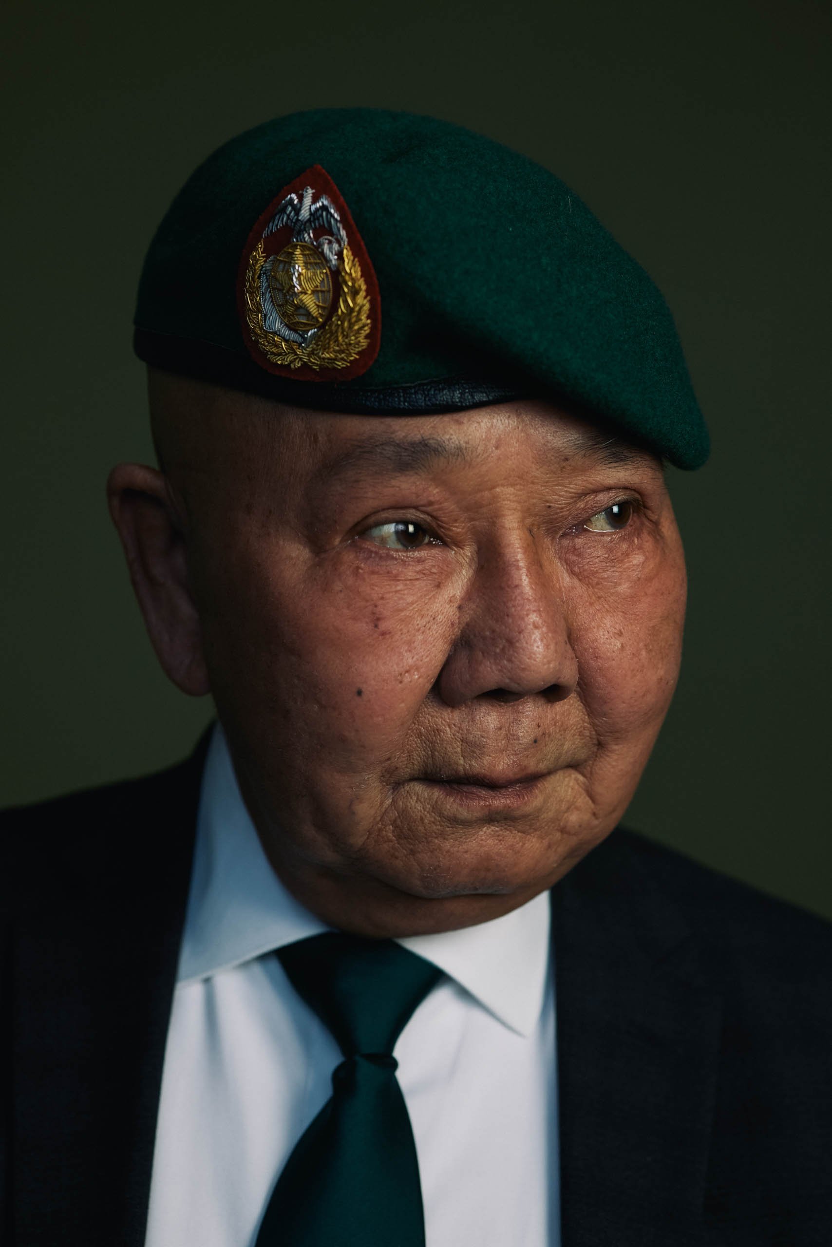 Portrait of an elderly man in a suit and green beret with a badge, gazing to the side against a green background in medium shot.