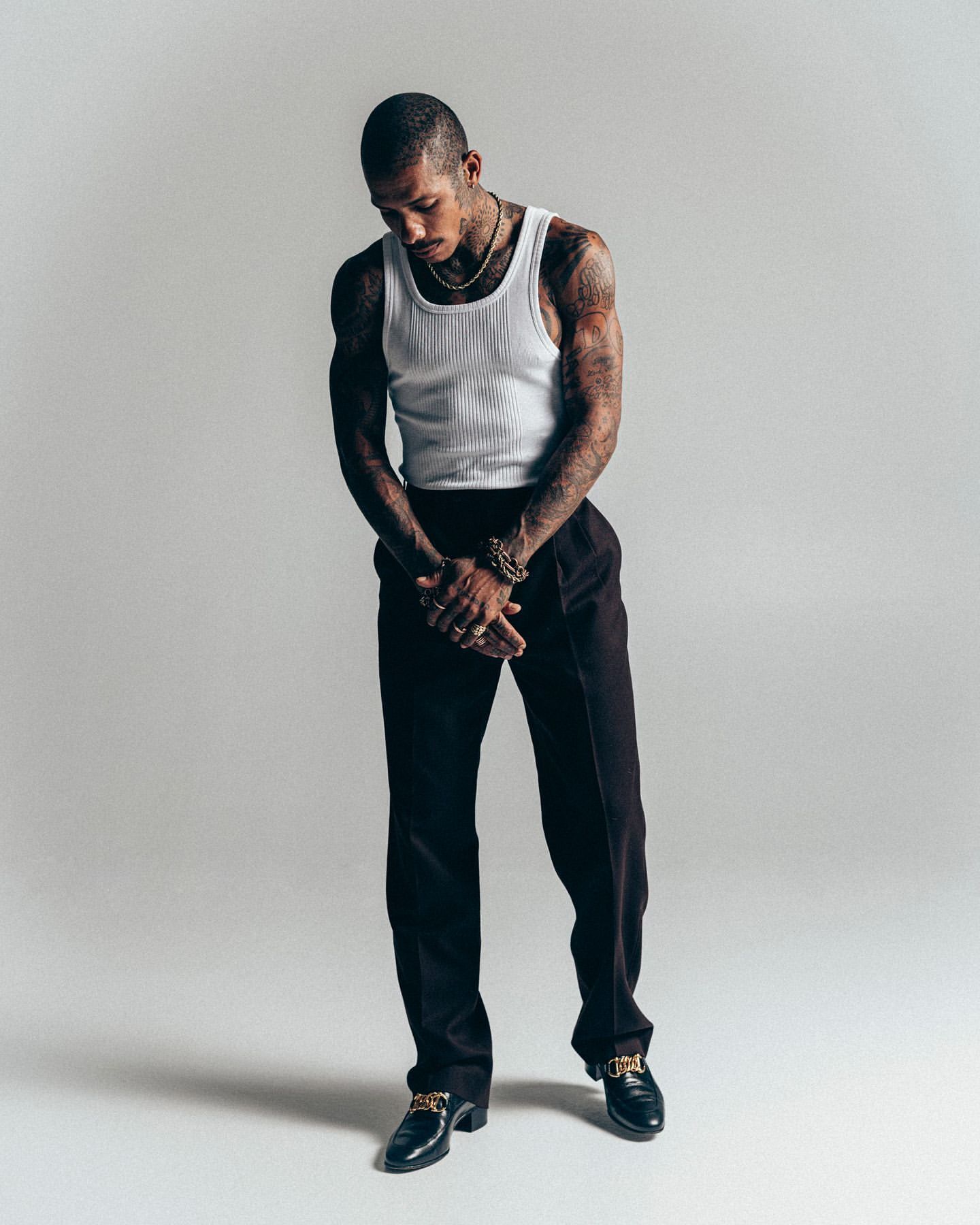Man with tattoos wearing a white tank top and dark trousers standing against a plain light grey studio background.