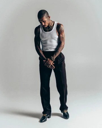 Man with tattoos wearing a white tank top and dark trousers standing against a plain light grey studio background.