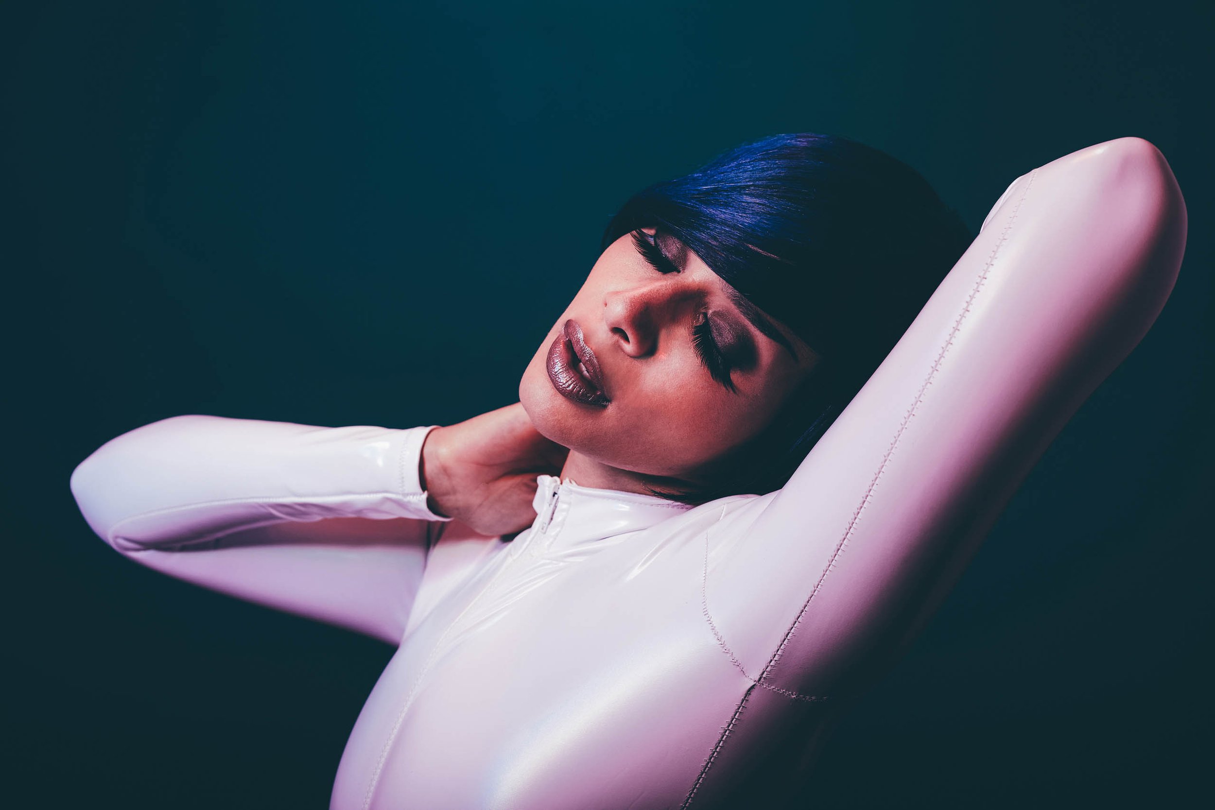 Portrait of a young adult woman in a white bodysuit against a dark backdrop, featuring a sleek hairstyle and dramatic lighting.
