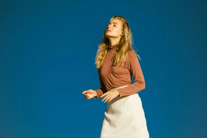 Blonde woman wearing a mauve turtleneck and white skirt against a vivid blue sky by Huy Doan