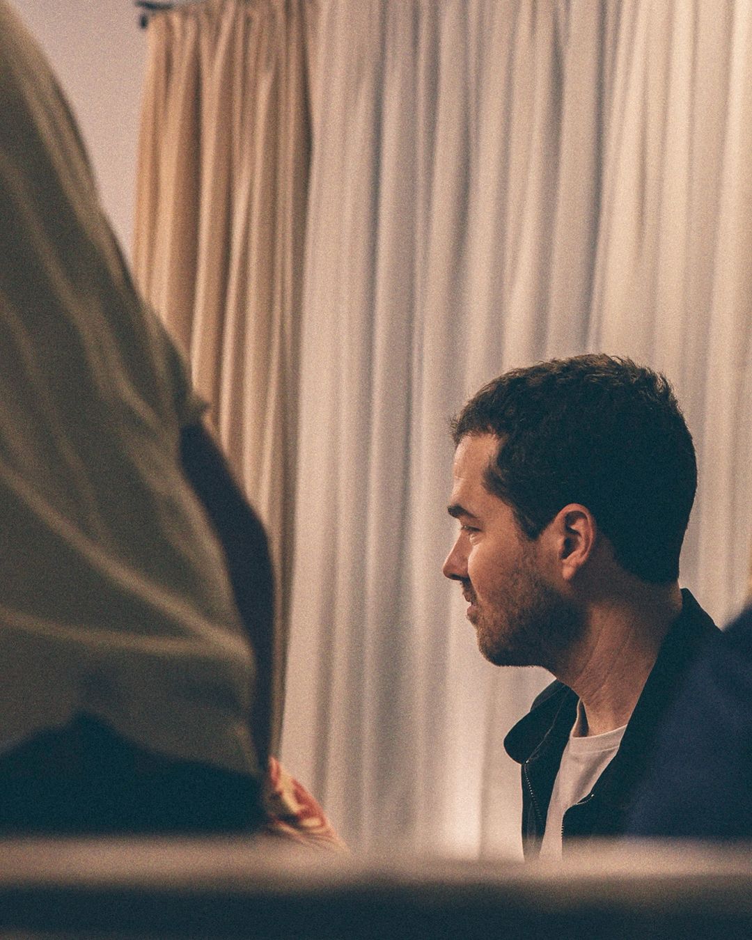 Man in navy jacket with a white t-shirt in a dimly lit room with a neutral-toned curtain in the background, looking toward another person.