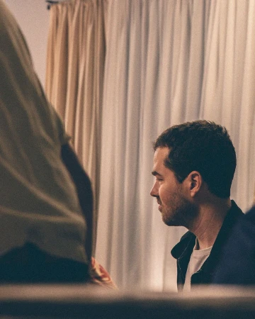Man in navy jacket with a white t-shirt in a dimly lit room with a neutral-toned curtain in the background, looking toward another person.