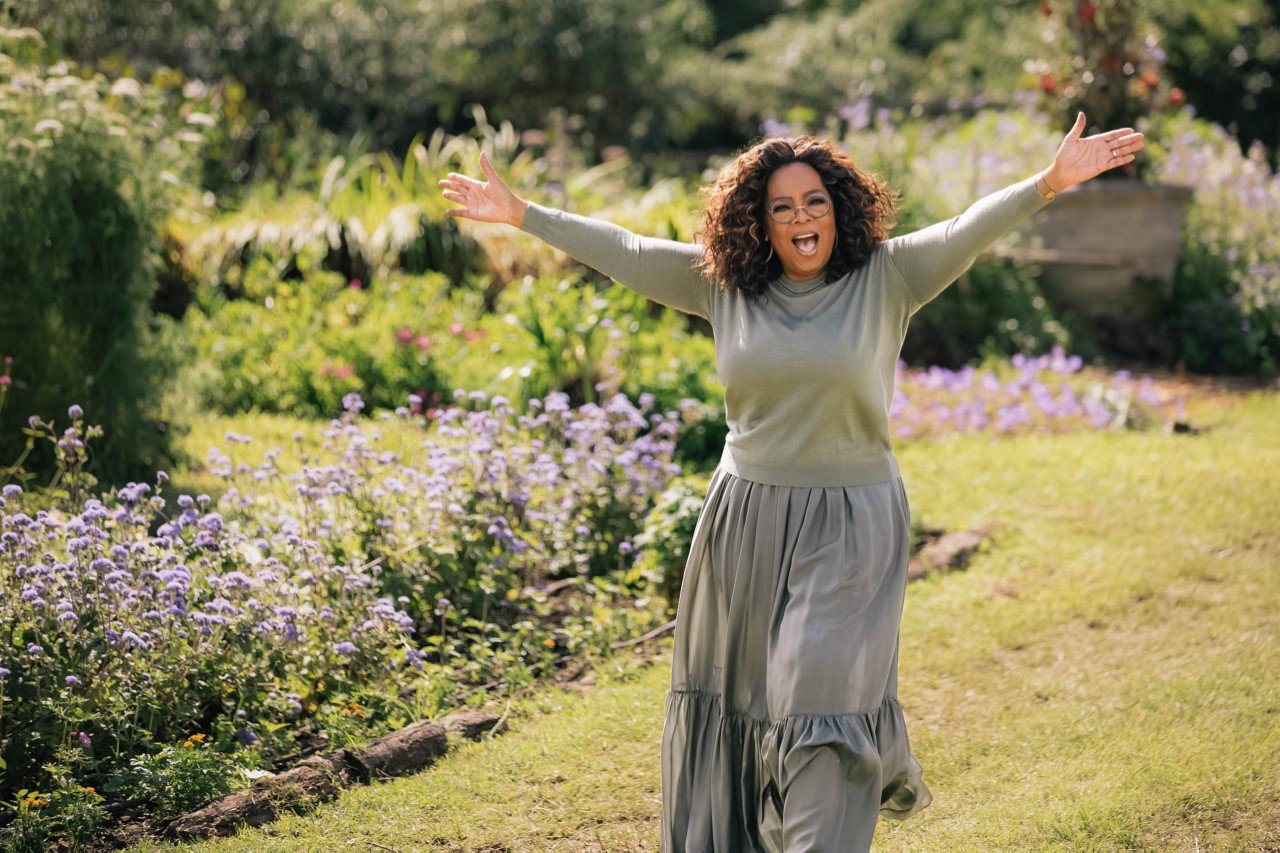 Oprah Winfrey stands in a lush garden with her arms outstretched in a welcoming gesture, wearing a casual green outfit.