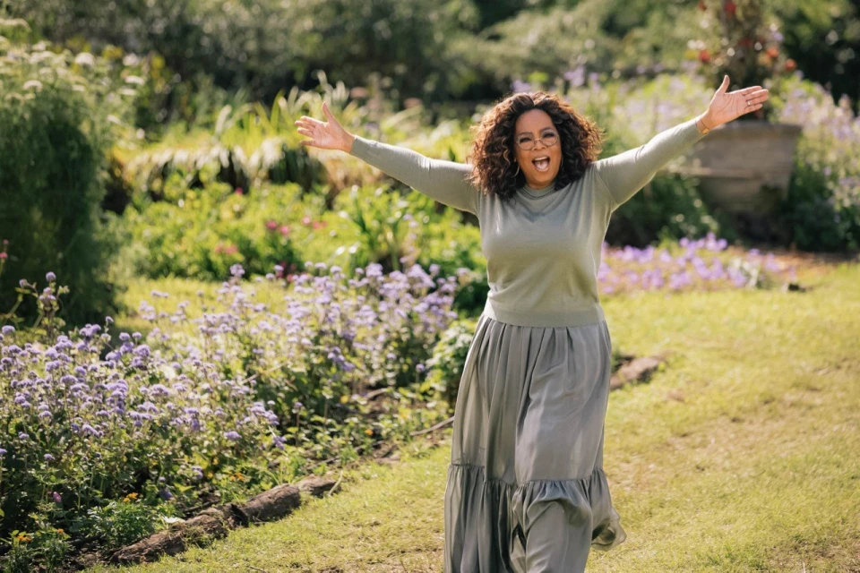 Oprah Winfrey stands in a lush garden with her arms outstretched in a welcoming gesture, wearing a casual green outfit.