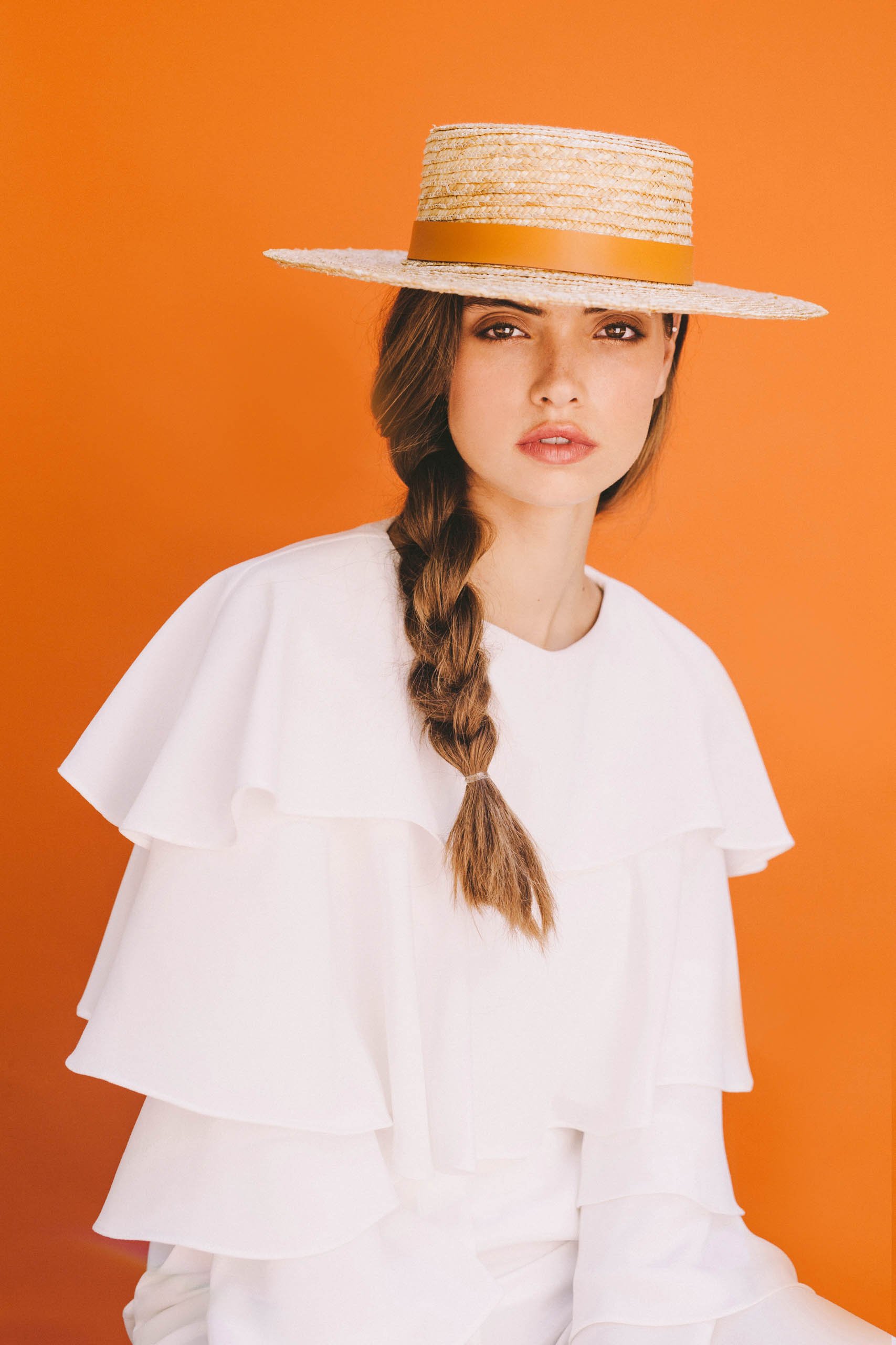 Portrait of a female model wearing a straw hat with a yellow band, white top, and braided hair against an orange background.