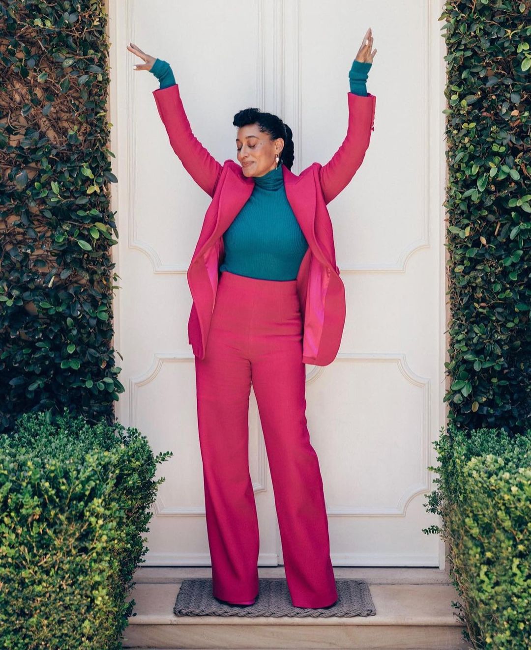 A full shot of a smiling woman with arms raised, wearing a bright pink suit and teal shirt, standing outside against a white door with green bushes.