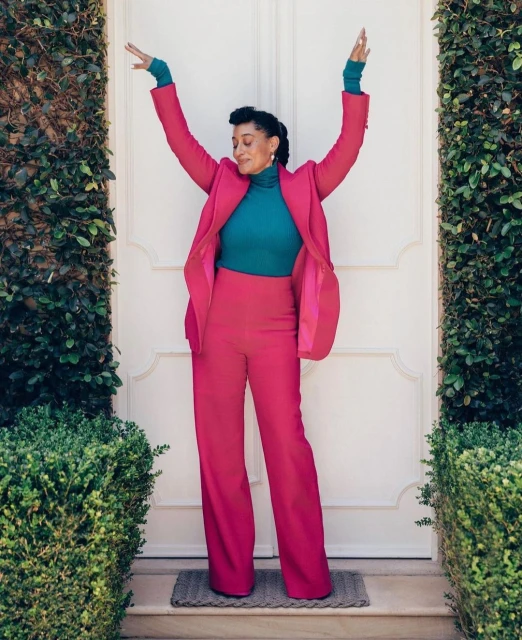 A full shot of a smiling woman with arms raised, wearing a bright pink suit and teal shirt, standing outside against a white door with green bushes.