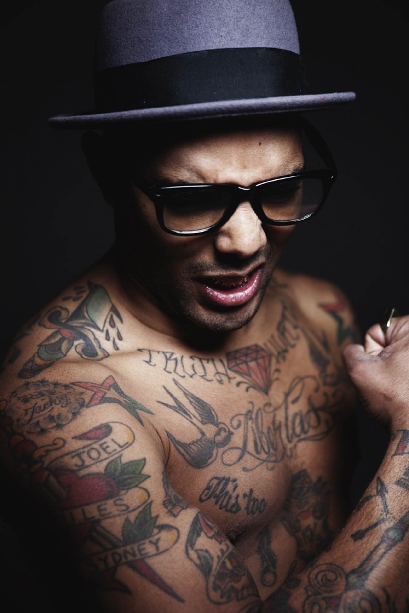 Portrait of a shirtless adult male wearing glasses and a hat with various tattoos against a dark backdrop in a photo studio.