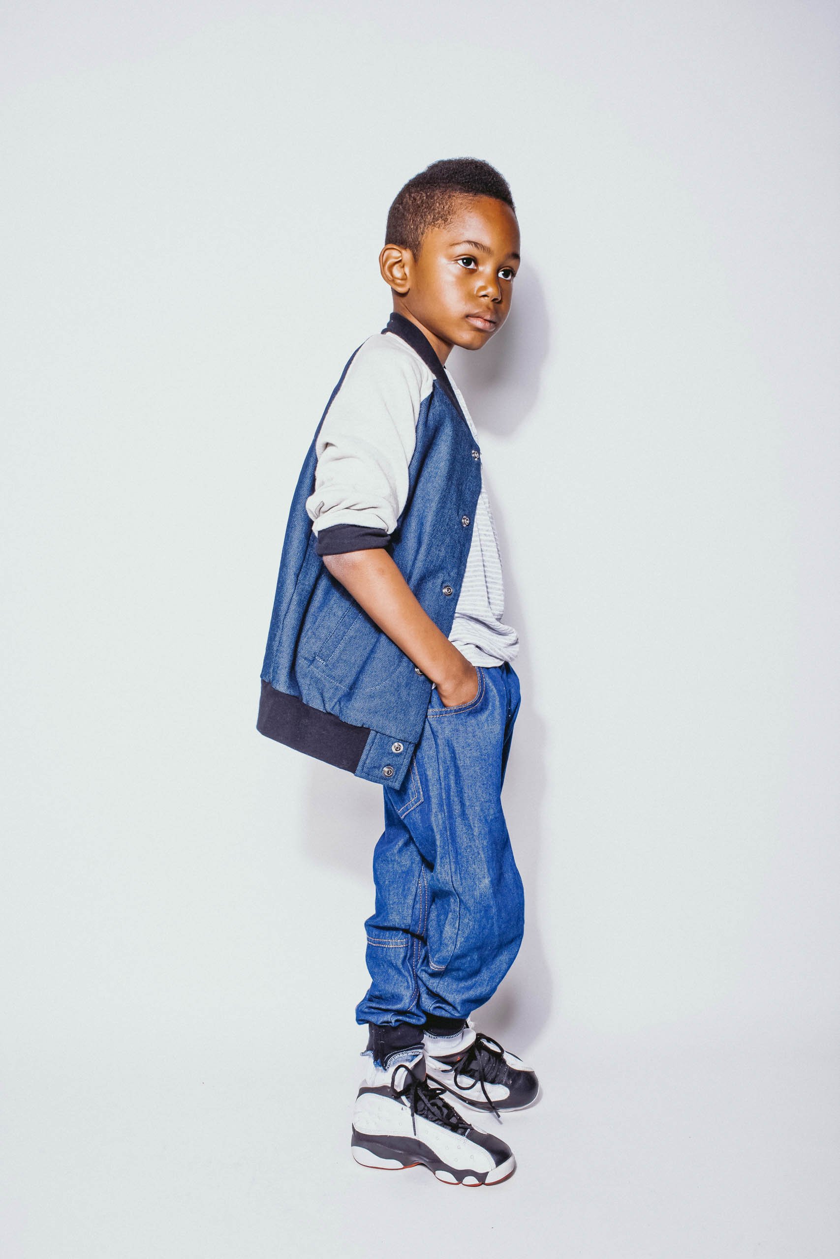 Young African-American boy in blue denim jacket and jeans stands indoors against a white backdrop in a fashion shot, looking away.