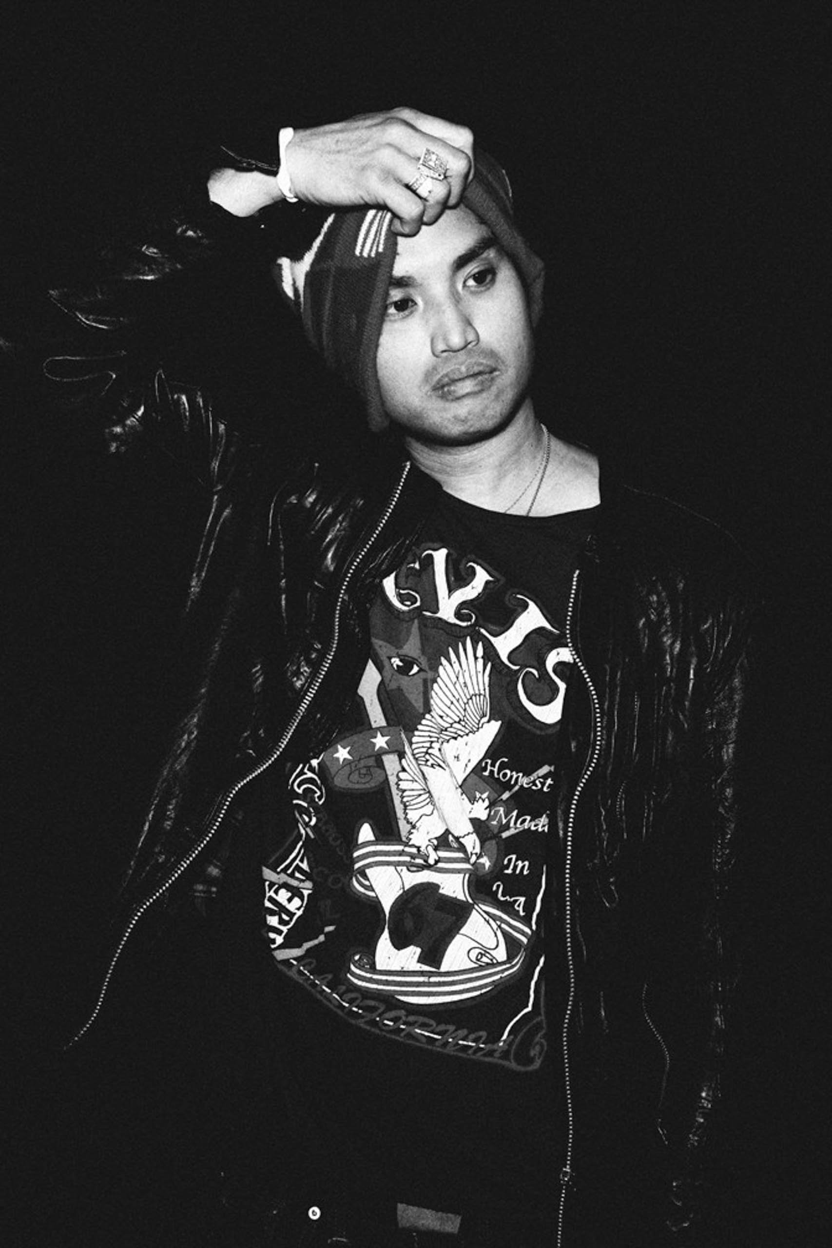Monochrome portrait of a man wearing a graphic t-shirt and dark jacket, adjusting his beanie against a black background.