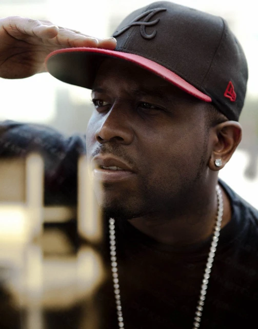 Close-up portrait of a man wearing a baseball cap with an 'A' logo and a beaded necklace. He is saluting with his hand.