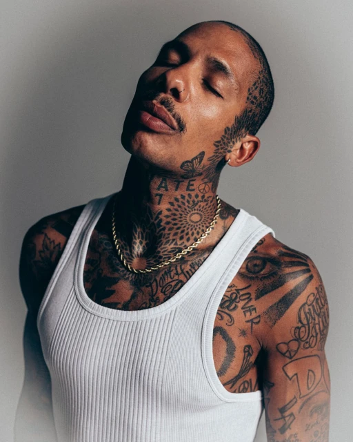 Portrait of a man with tattoos in a white ribbed tank top, eyes closed, featuring a gold chain against a neutral grey backdrop.